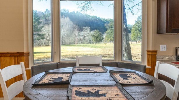 A round dining table with six chairs, a view of a grassy field and trees through large windows, and wooden paneling on the walls. - Riverfront country setting (Bath County)