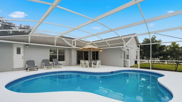 A covered swimming pool with lounge chairs and a table, surrounded by a white fence and trees. - Disney Marvelous 4-bedroom house with a pool (Kissimmee)