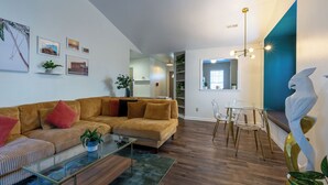 A modern living room with a sofa, coffee table, and chairs. There are framed pictures on the wall and a plant on the floor. - Stylish Durham Retreat • 3BR, Private Backyard • Near NCCU, Duke & Downtown (Durham)