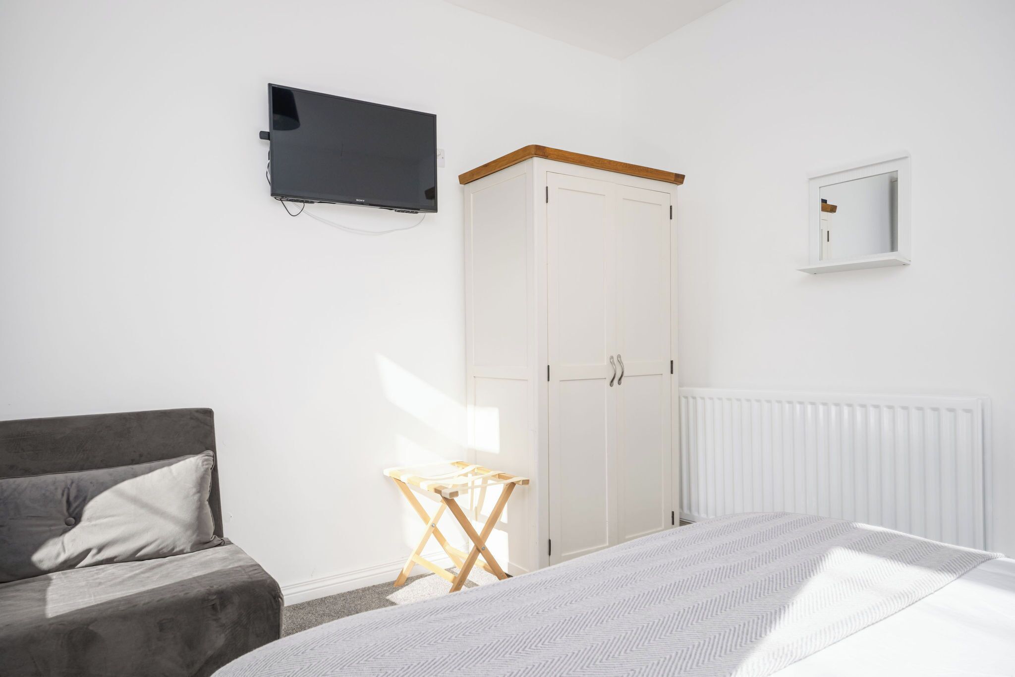 Une chambre à coucher avec un lit, un canapé gris, une armoire, une petite table et un téléviseur fixé au mur.