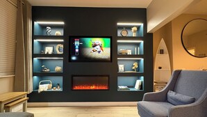 Una sala moderna con un televisor de pantalla plana montado en una pared oscura, rodeada de estanterías con objetos decorativos y una chimenea abajo.