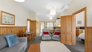 Una habitación acogedora con un sofá gris, una mesa de comedor de madera, dos sillones, una alfombra pequeña y una cocina visible a través de una puerta abierta.