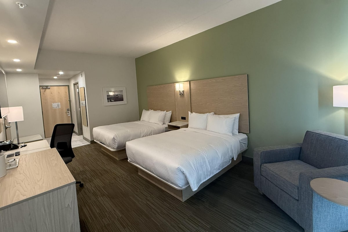 A hotel room with two beds, a desk, a chair, a sofa, and a wall-mounted lamp.