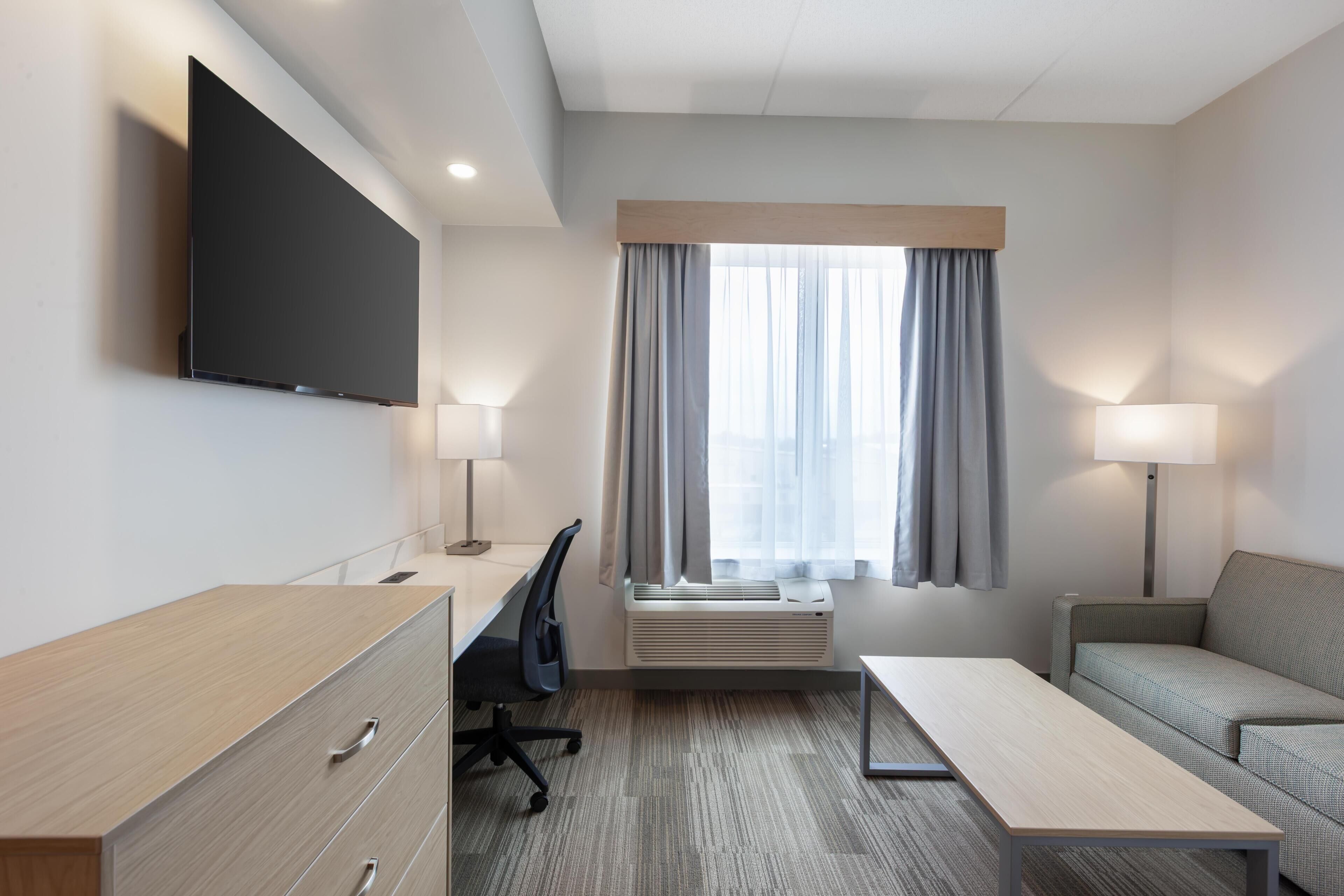 A hotel room with a flat-screen TV, a desk with a chair, a sofa, two lamps, and a large window with curtains.