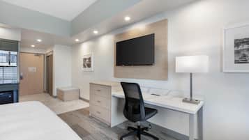 A modern hotel room with a bed, desk, chair, TV, and artwork on the walls.