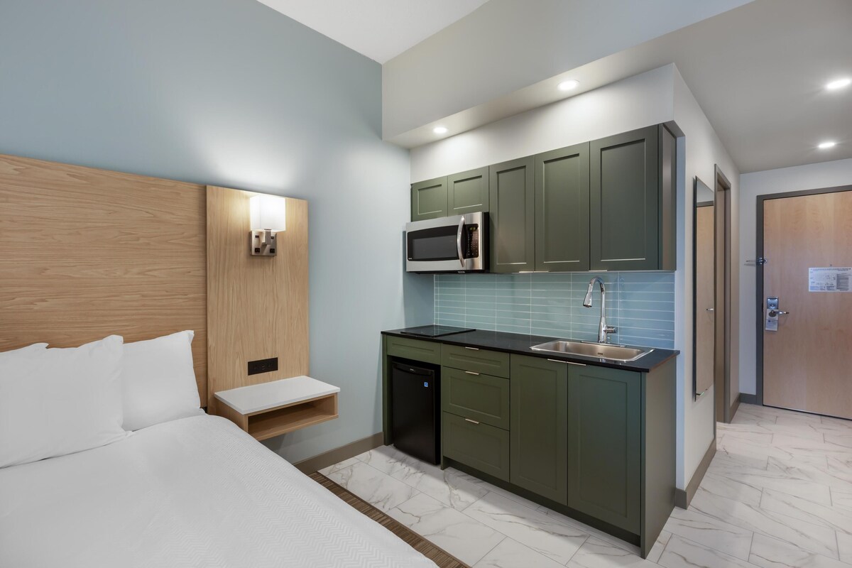 Een compacte hotelkamer met een bed, een klein nachtkastje, een ingebouwde magnetron, een wastafel en een compacte kitchenette.