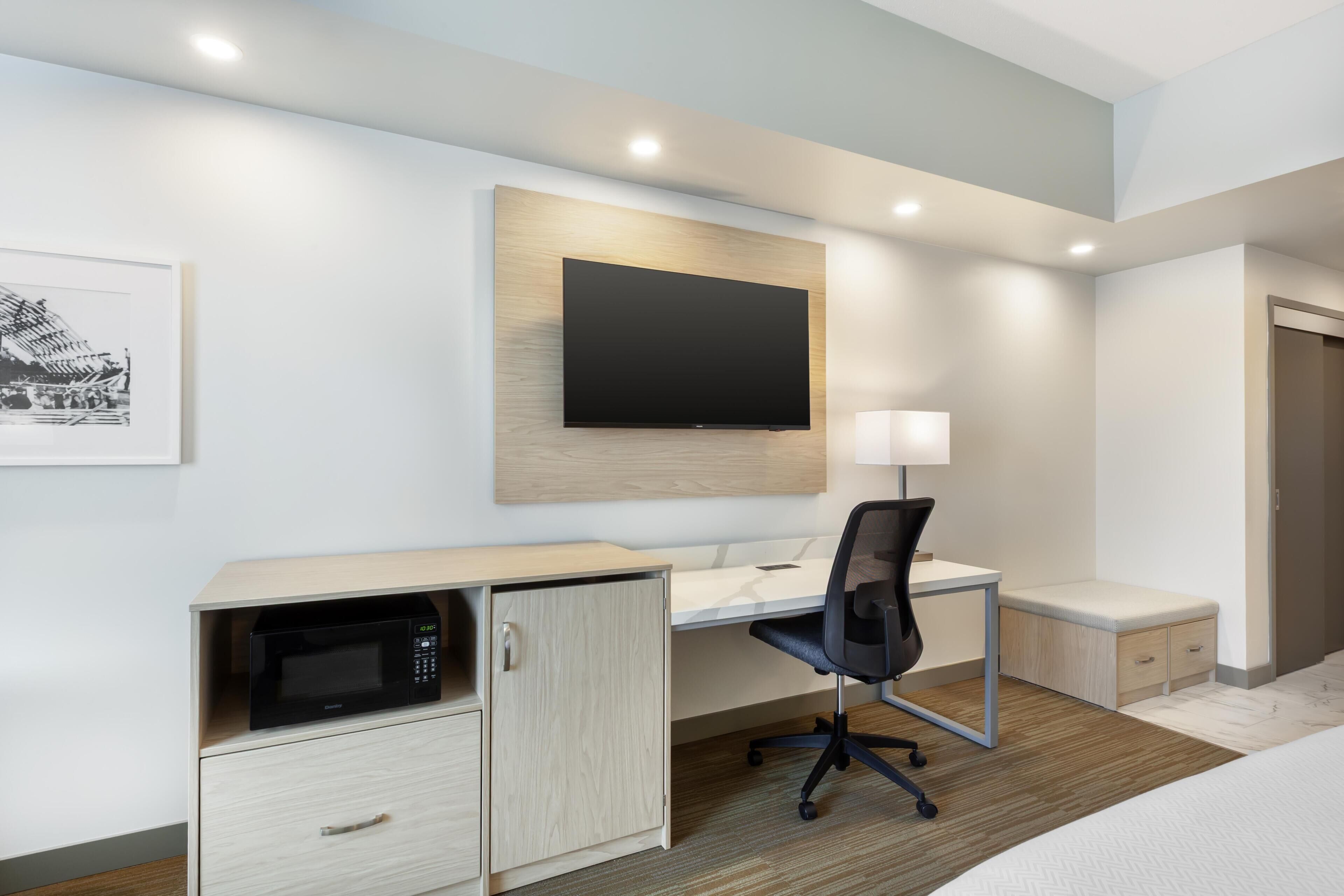 A hotel room with a bed, a desk with a chair, a microwave, a television, a bedside lamp, and a door.