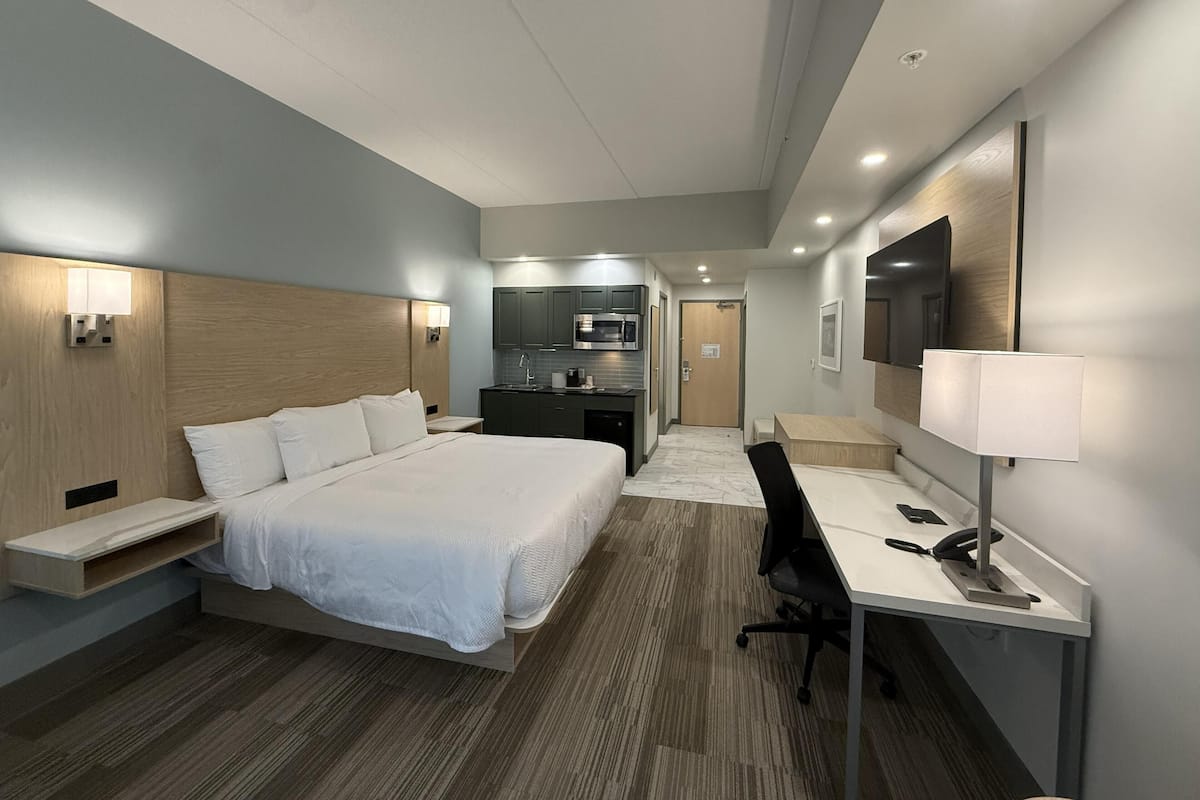 A hotel room with a bed, desk, chair, kitchenette, and a TV.