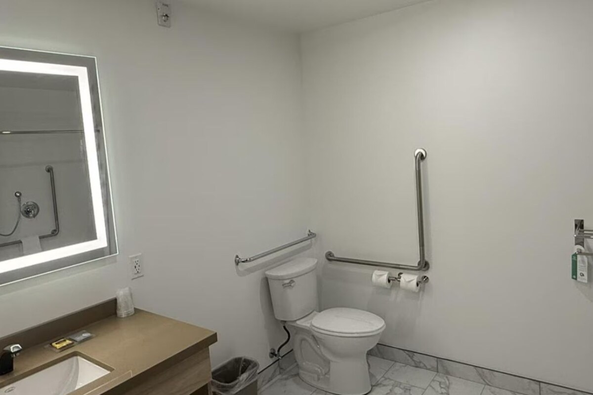 Une salle de bain avec un grand miroir, des toilettes équipées d’une barre d’appui, un lavabo et une poubelle.