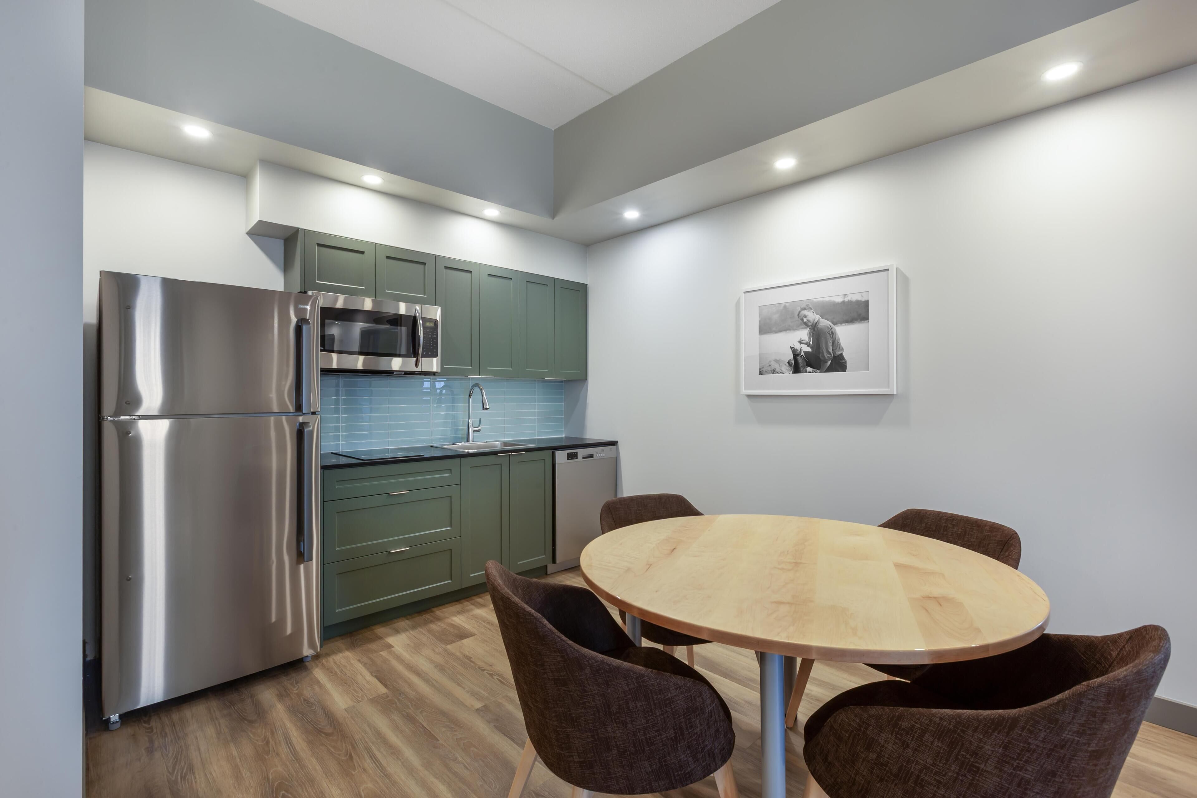 A modern kitchen with a stainless steel refrigerator, microwave, and dishwasher, green cabinets, a wooden dining table with four chairs, and a framed picture on the wall.