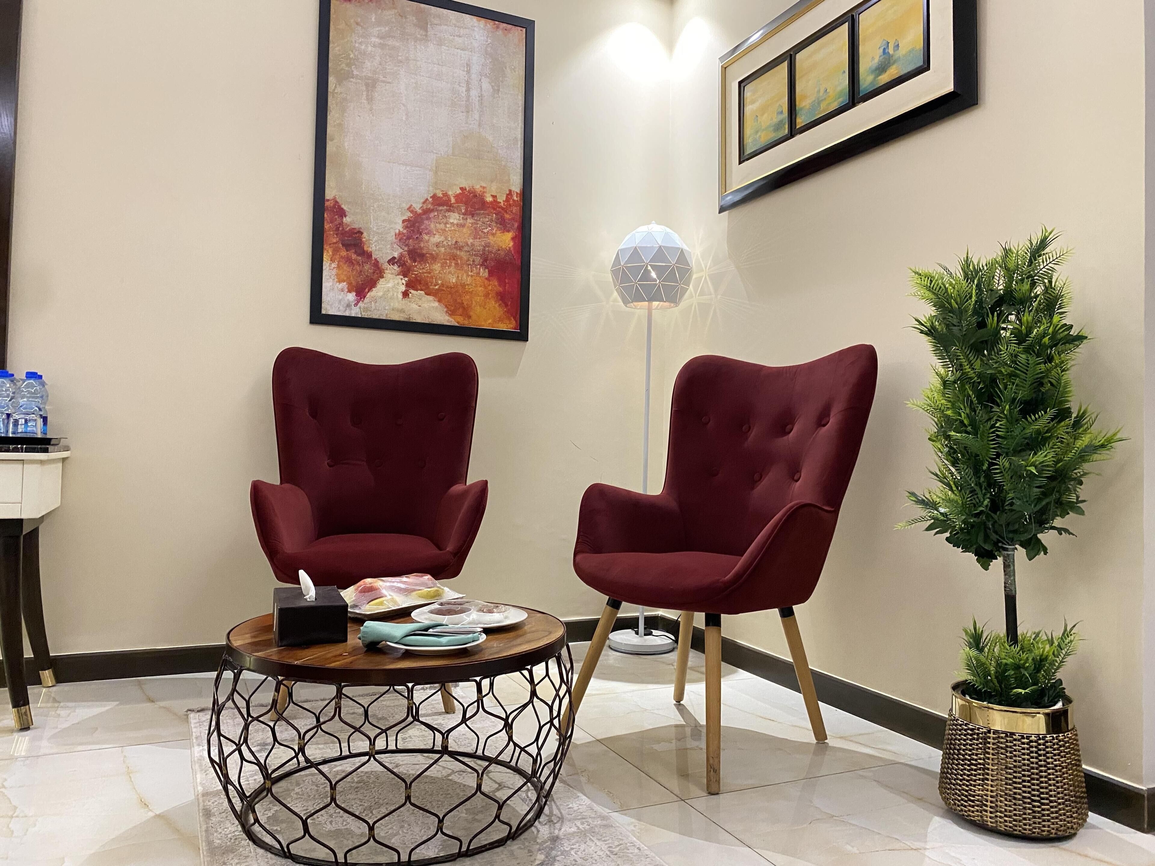 Un vestíbulo moderno con dos sillones rojos, una mesa redonda de base metálica geométrica, una lámpara de pie y una planta en maceta.