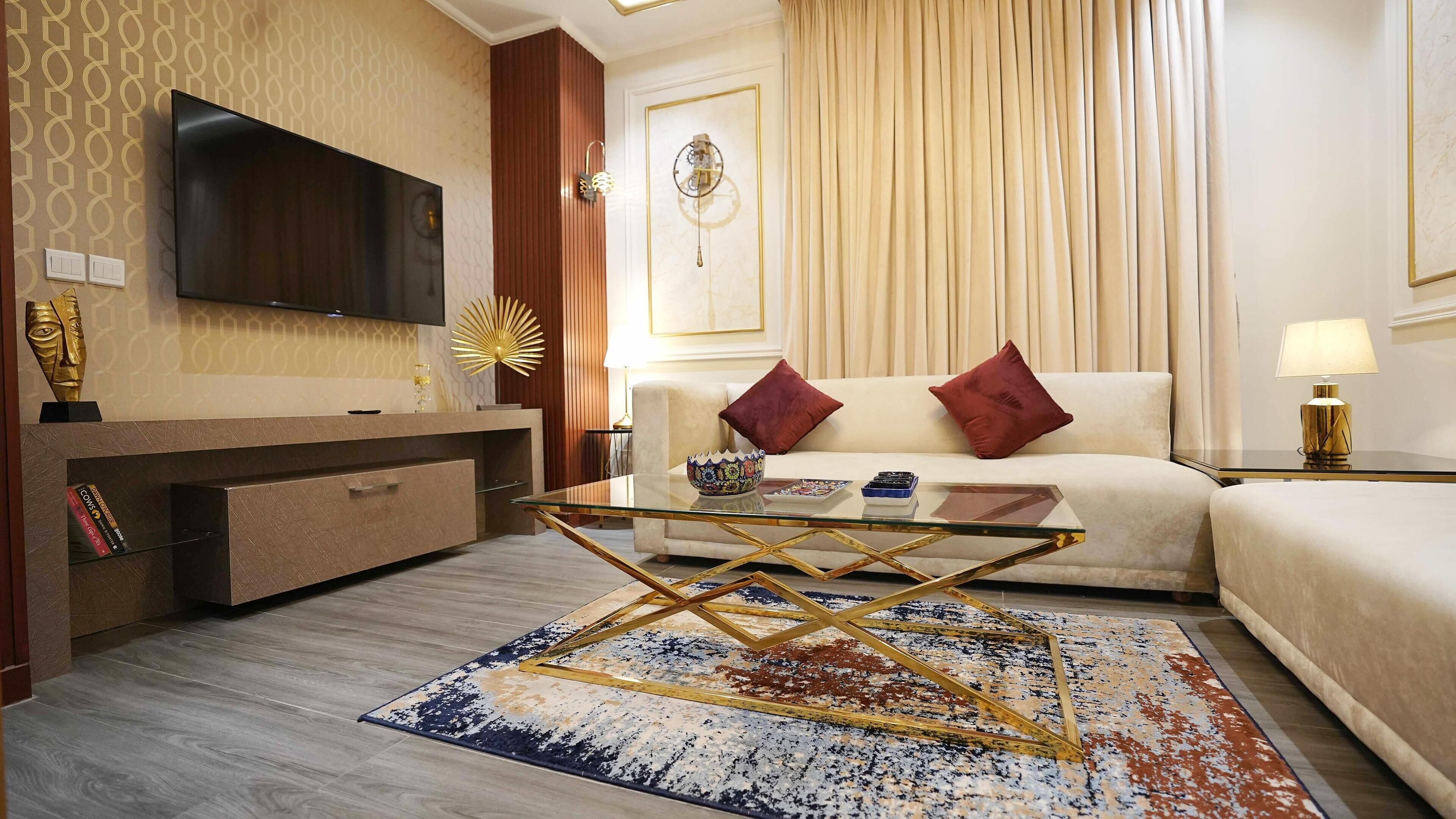 Una habitación de hotel moderna con una televisión de pantalla plana, una mesa de centro de vidrio, un sofá con cojines rojos, un elemento decorativo en la pared y una alfombra estampada.