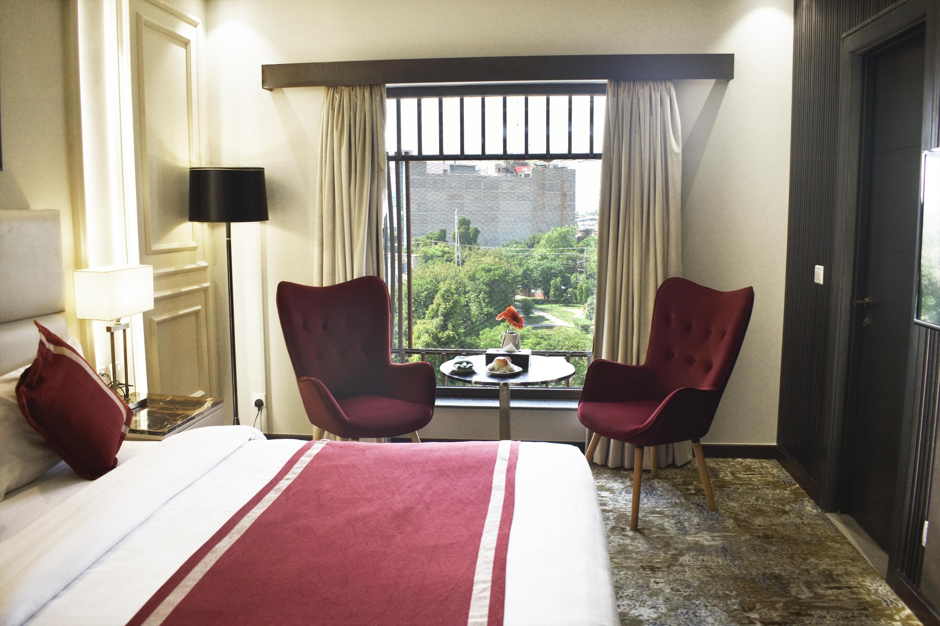 Habitación de hotel con cama, dos sillas rojas, una mesita con un florero y ventana con vistas a zonas verdes y edificios.