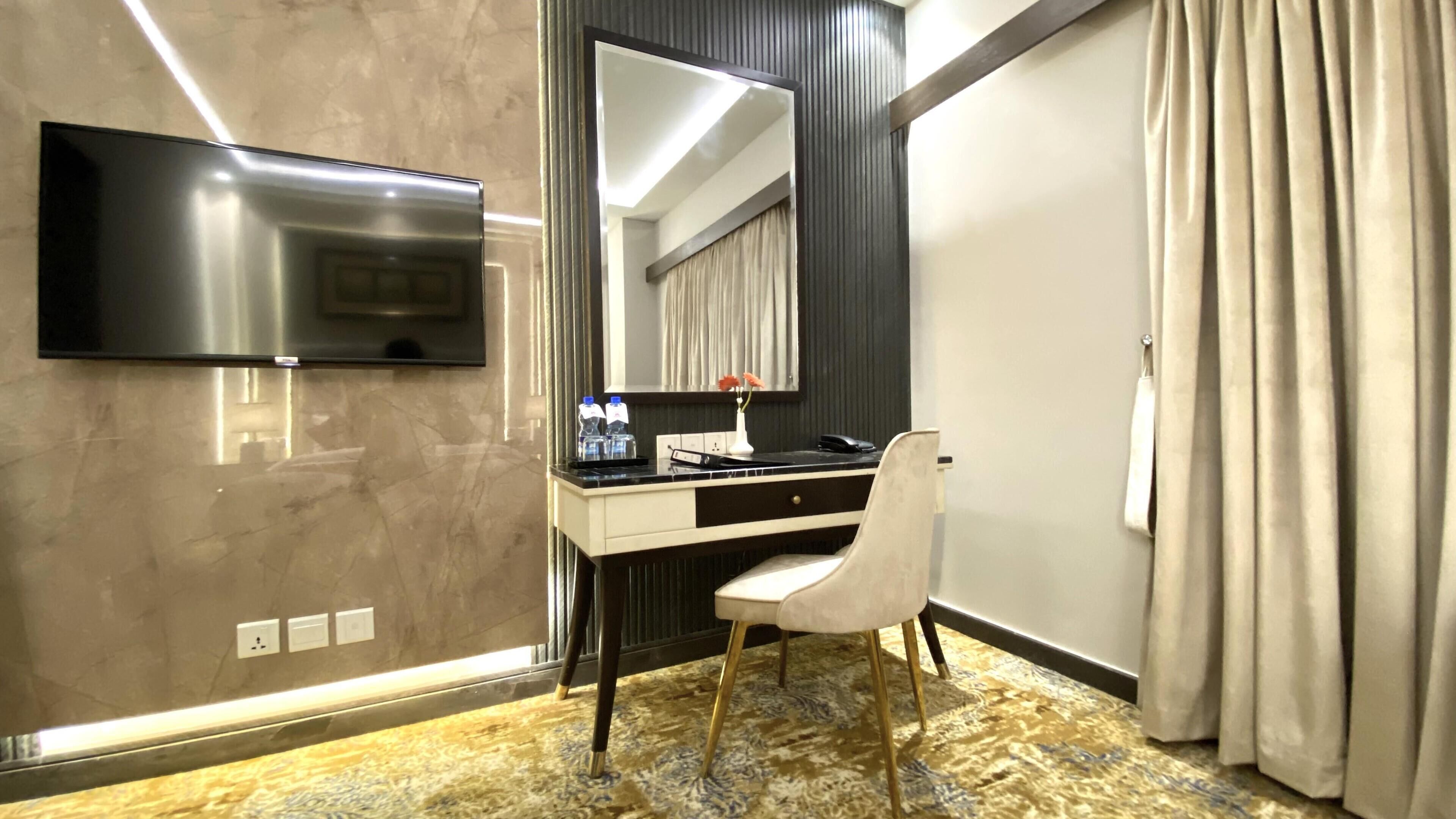 A hotel room with a modern desk, a flat-screen TV mounted on the wall, a large mirror, and a bed with curtains.