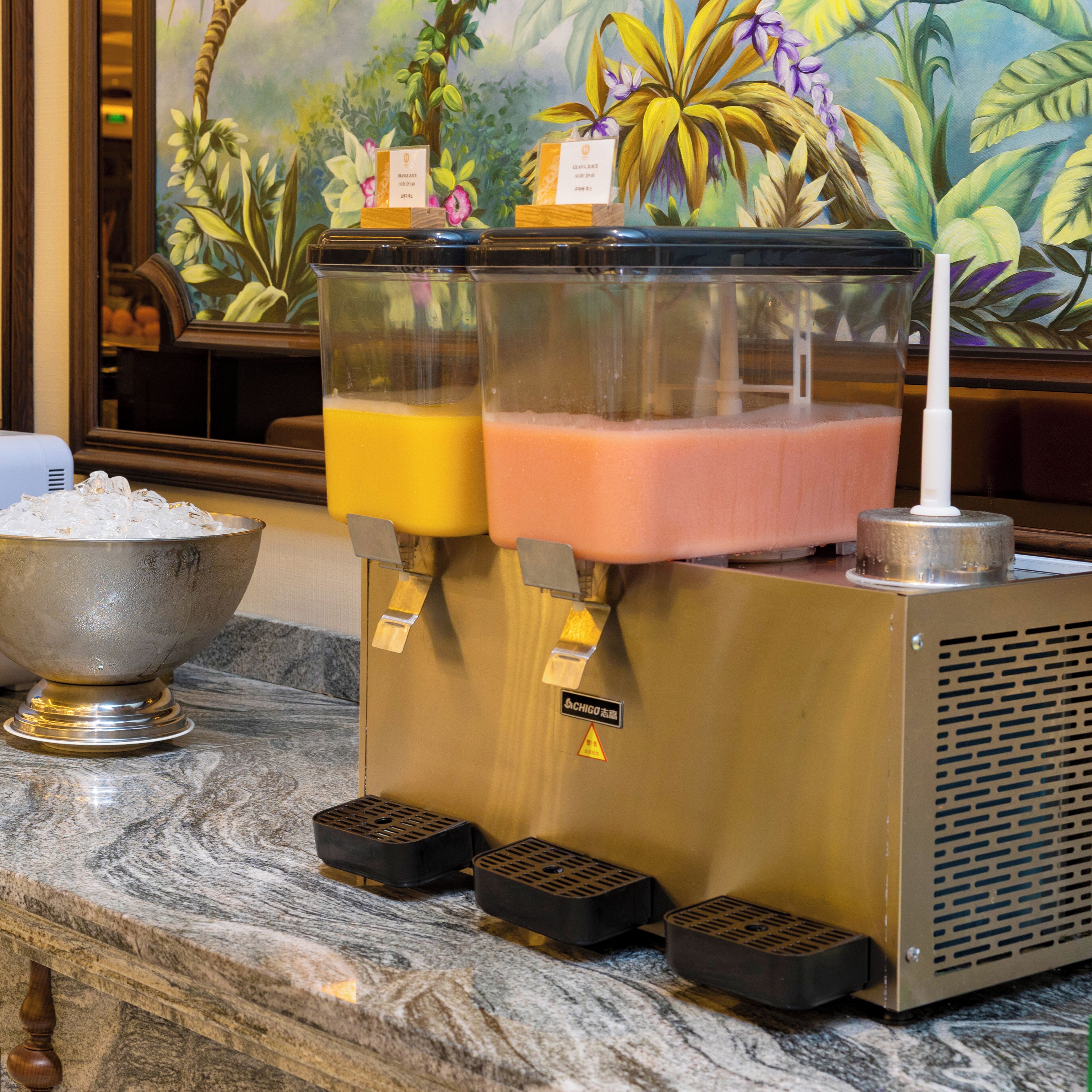 A juice dispenser with two containers, a bowl of ice, and a decorative mural in the background.