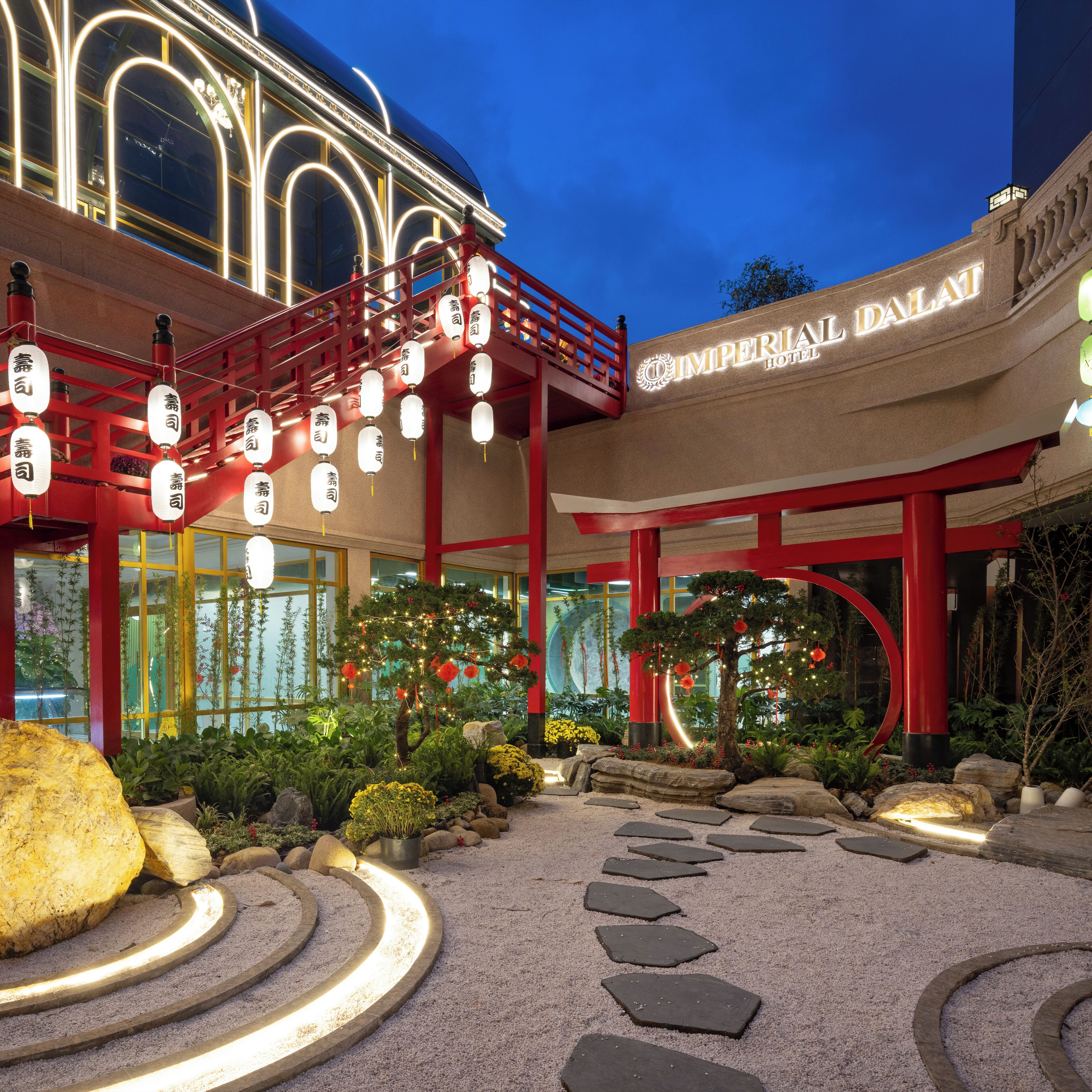 Fachada roja y blanca de un hotel, faroles tradicionales y un jardín ajardinado con piedras.