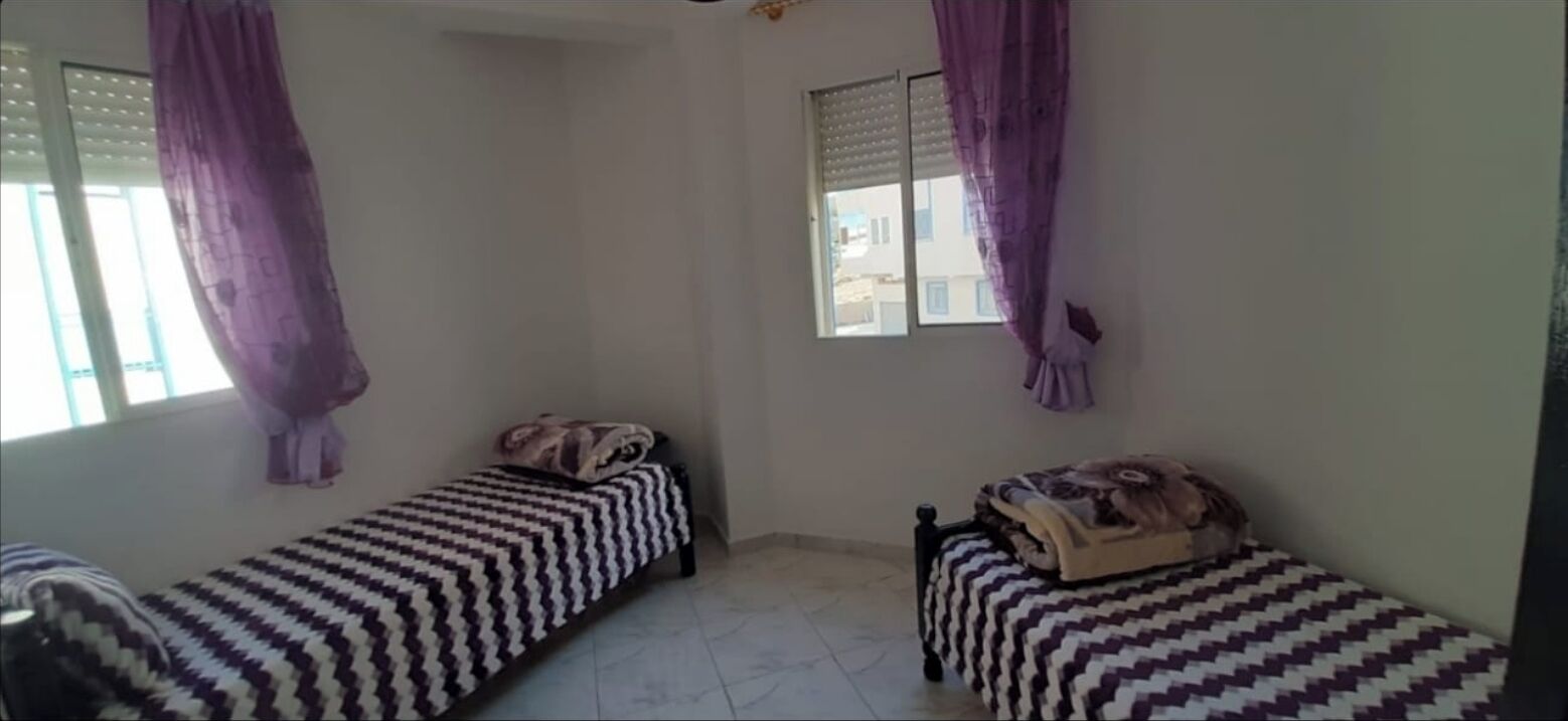 A small, single-room with two single beds, each with patterned bedding. There is a window with purple curtains, and the room has plain walls and a tiled floor.