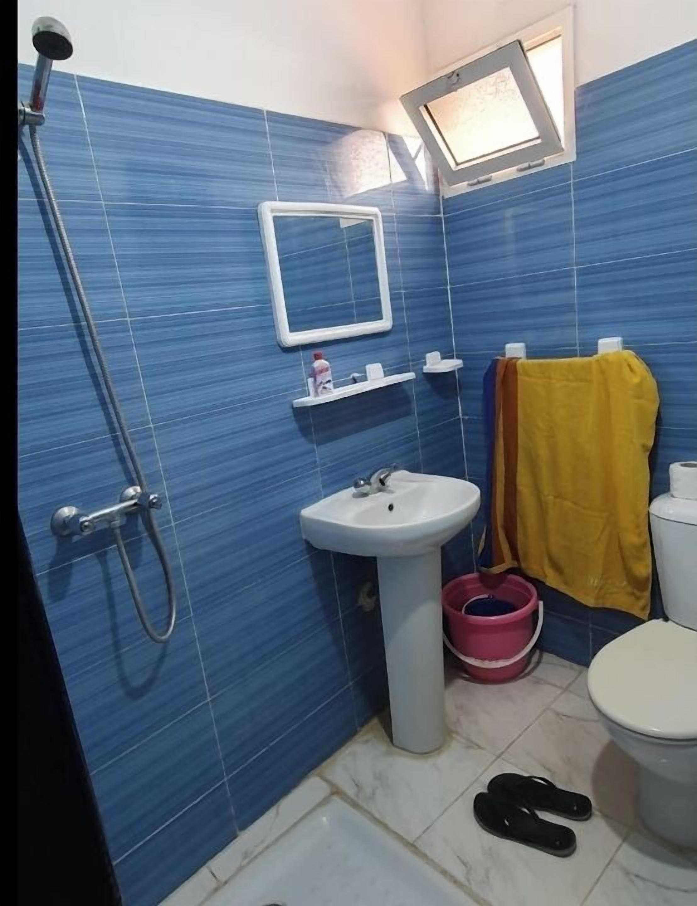 Un baño con azulejos azules, un lavamanos blanco, un espejo, una toalla amarilla, un cubo rosa y un inodoro.