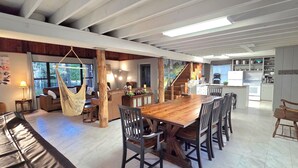 Ein großzügiger Wohnbereich mit einem großen Esstisch aus Holz, bequemen Sitzgelegenheiten, einem Hängekorbstuhl und einer Küche im Hintergrund.