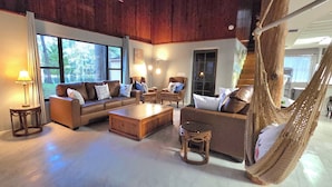 Ein großzügiges Wohnzimmer mit einer großen Sofagarnitur, einem Couchtisch und einer Hängematte. Mehrere Fenster sorgen für natürliches Licht und einen Blick ins Freie.
