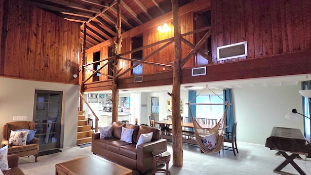 Ein großzügiger Wohnbereich mit hoher Decke, Holzbalken, einer zentralen Sitzecke mit Sofa und Sesseln, einem Hängesessel und einer Treppe, die in einen oberen Bereich führt.