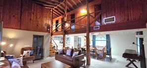 A spacious living area with a high ceiling, wooden beams, a central seating area with a sofa and chairs, a hanging chair, and a staircase leading to an upper level. - Rainbow River! Kayaks/Tubing/Sauna Sleeps 12 
Triple R Retreat Rest-Relax-Repeat (Dunnellon)