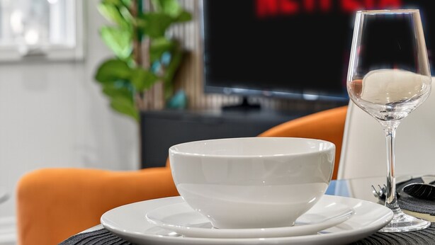 A dining area with a table set for two, a glass of water, and a bowl. A television with the Netflix logo is in the background. - Worldcup Ready Stylish 1BR in West Village  Pool + Free Parking (Dallas)