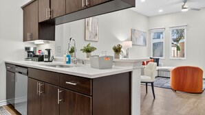 A modern kitchen with dark wood cabinets, a white countertop, and stainless steel appliances. There is a dining area with a white chair and a colorful orange chair. - Worldcup Ready Stylish 1BR in West Village  Pool + Free Parking (Dallas)