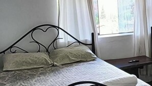 Una camera da letto con un letto, due cuscini, un comodino, una finestra con tende e un testiera a forma di cuore in metallo.