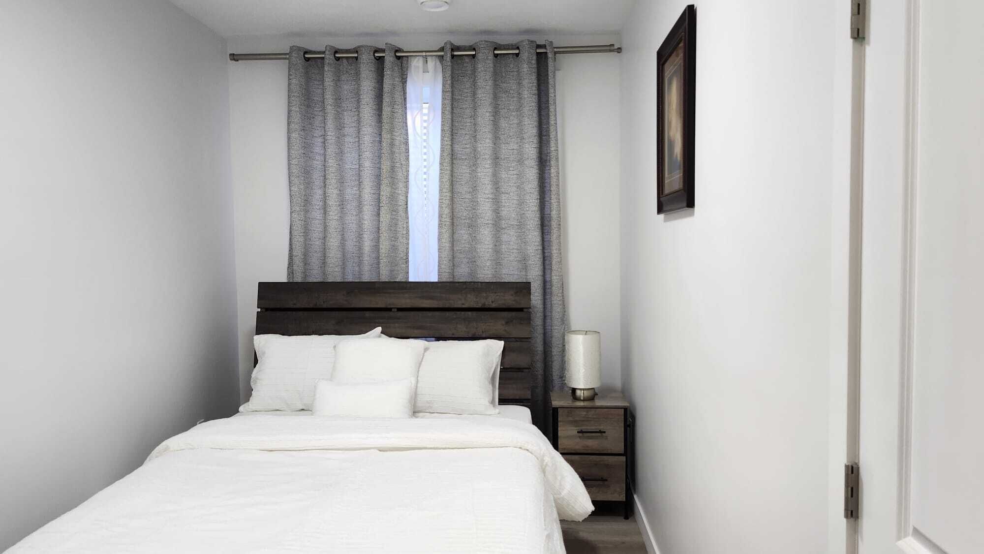 Une chambre à coucher avec une tête de lit en bois, du linge de lit blanc, une table de chevet avec une lampe et une fenêtre avec des rideaux gris.