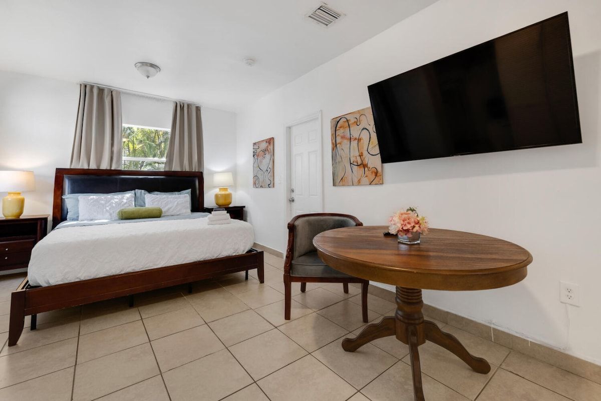 Une chambre d’hôtel avec un lit, une table ronde en bois, une chaise, un téléviseur fixé au mur et des œuvres d’art accrochées aux murs.