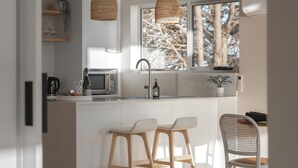 A modern kitchen with white cabinetry, a central island with stools, a microwave, and a hanging woven light fixture. - Couples Luxury Retreat with Ocean Views and Outdoor Bath (Penguin)