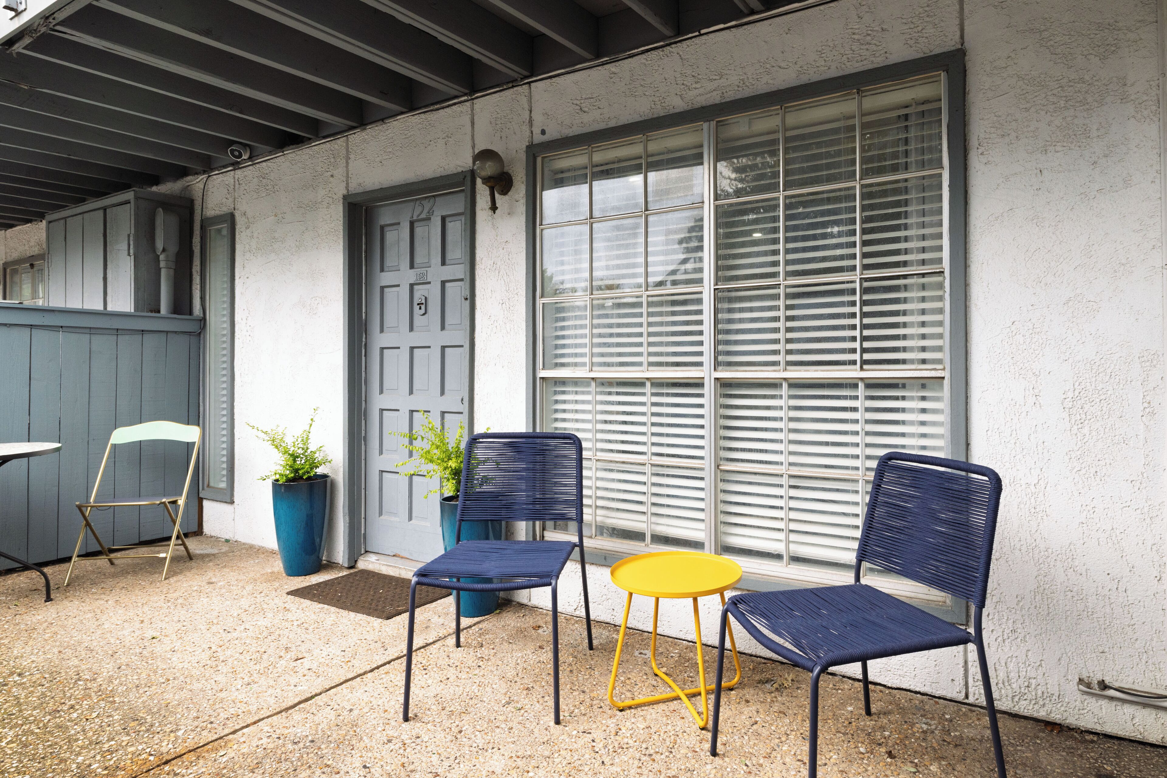 A patio area with two blue chairs and a yellow table, a potted plant, and a door with a number 101 sign.