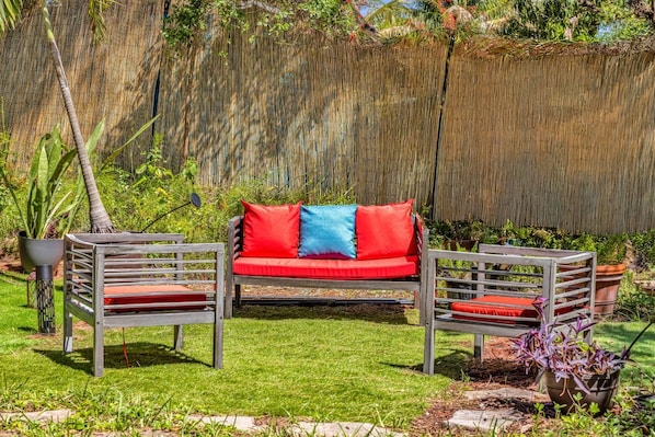 Un jardin avec un canapé rouge et deux fauteuils, une clôture en bambou et des plantes en pot.
