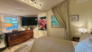 Habitación de hotel con cama, cómoda, televisión, un cuadro, ventana con cortinas y mesita de noche con lámpara.