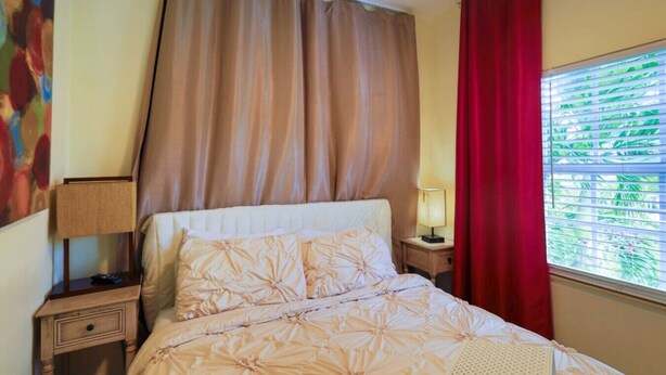 A hotel room with a bed, a nightstand, a lamp, a window with blinds, and a painting on the wall. - Suarez Art Stay, 3BR Miami Oasis with Parking (Miami)