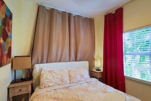 A hotel room with a bed, a nightstand, a lamp, a window with blinds, and a painting on the wall. - Suarez Art Stay, 3BR Miami Oasis with Parking (Miami)