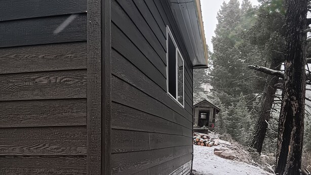 Ein Holzhaus mit Fenster, umgeben von schneebedecktem Boden und Bäumen.