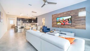 A modern living room with a large flat-screen TV mounted on a wooden wall, a ceiling fan, and a white sofa with orange cushions. - Go 02 Champions Gate (Davenport)