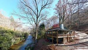 A cabin with a deck overlooking a river, surrounded by trees and a forested area. - Steps to Shooting Creek (Hayesville)