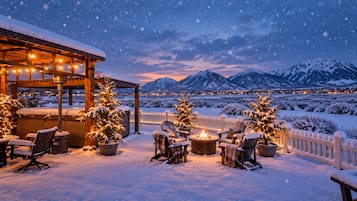 A snow-covered outdoor seating area with chairs and a fire pit, surrounded by snow-covered trees and mountains in the background.