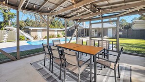 A covered patio with a pool table and chairs, overlooking a backyard with a pool and a slide.