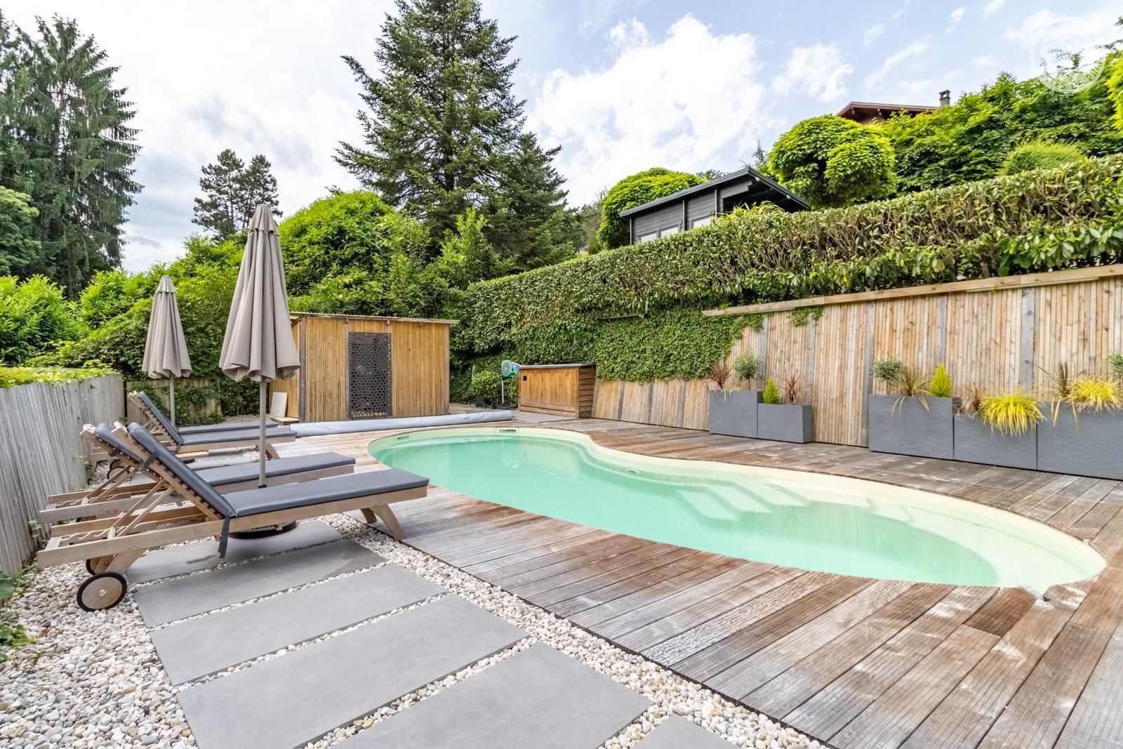Una piscina con sillas de descanso y sombrillas, rodeada por una terraza de madera y una valla de madera.