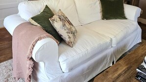 En hvid sofa med flere puder, et træ sofabord, et spejl og en hængende lampe i en stue med beige vægge og trægulv.
