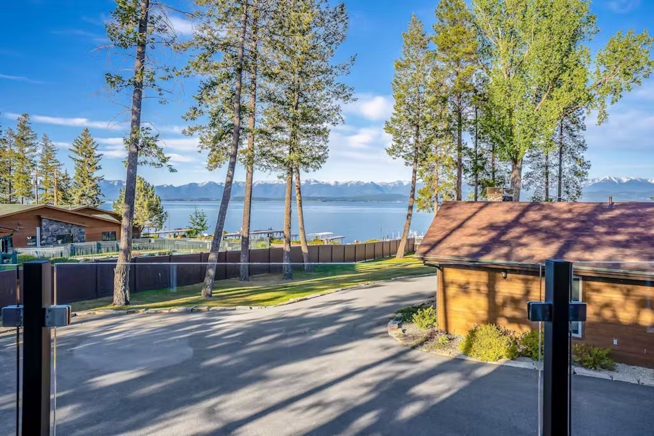 A view of a road leading to a building with a wooden roof, surrounded by tall trees and a body of water with mountains in the background.