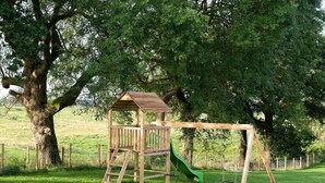 Una estructura de joc de fusta amb tobogan i cadira de balanceig en una zona verda amb gespa.