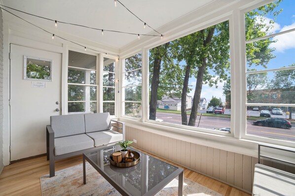 Ein Zimmer mit einer Couch, einem Glastisch und großen Fenstern mit Blick auf die Straße und Bäume.