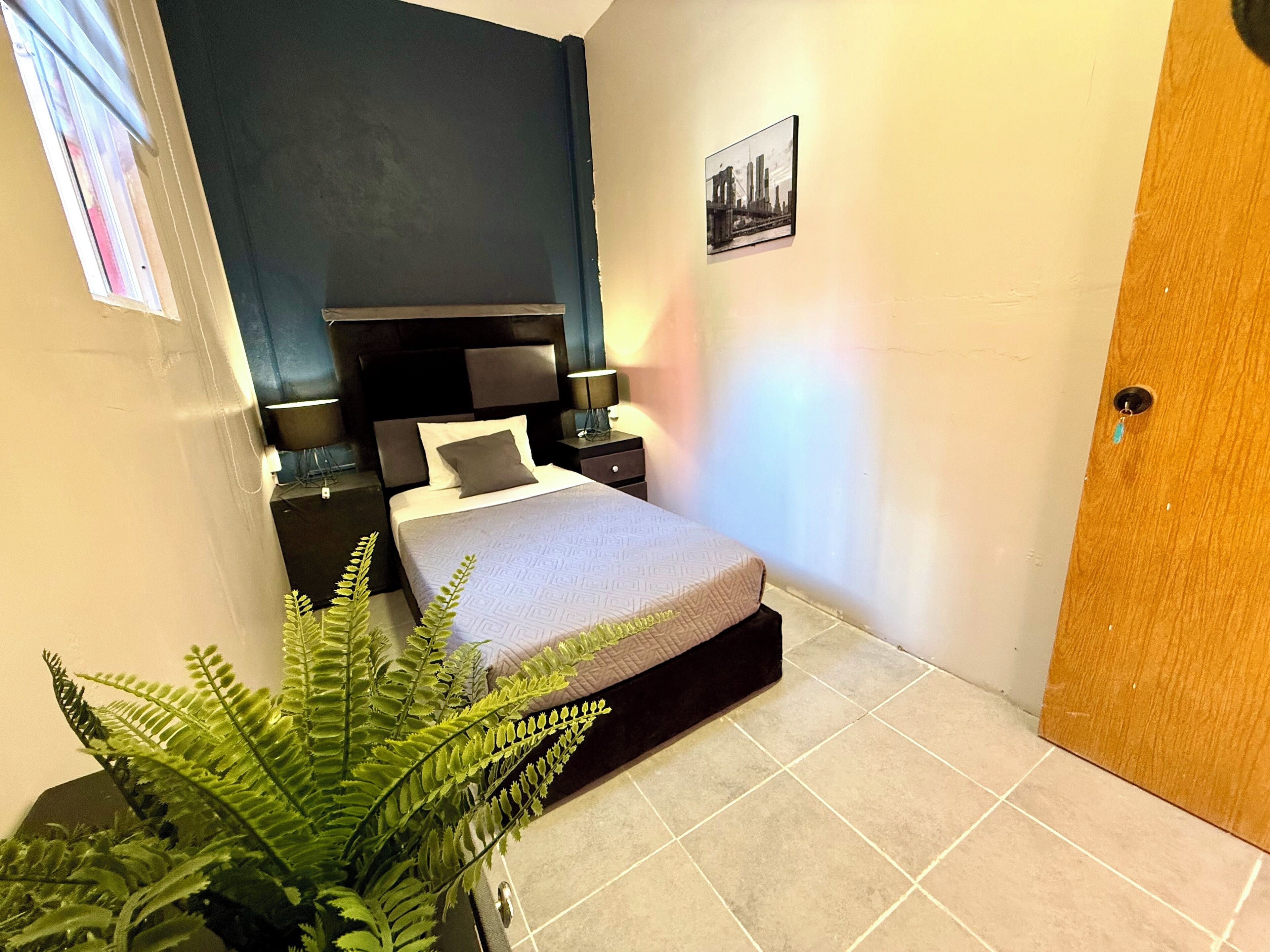 A small, neatly arranged bedroom with a bed, bedside tables, a nightstand, a potted plant, and a door.
