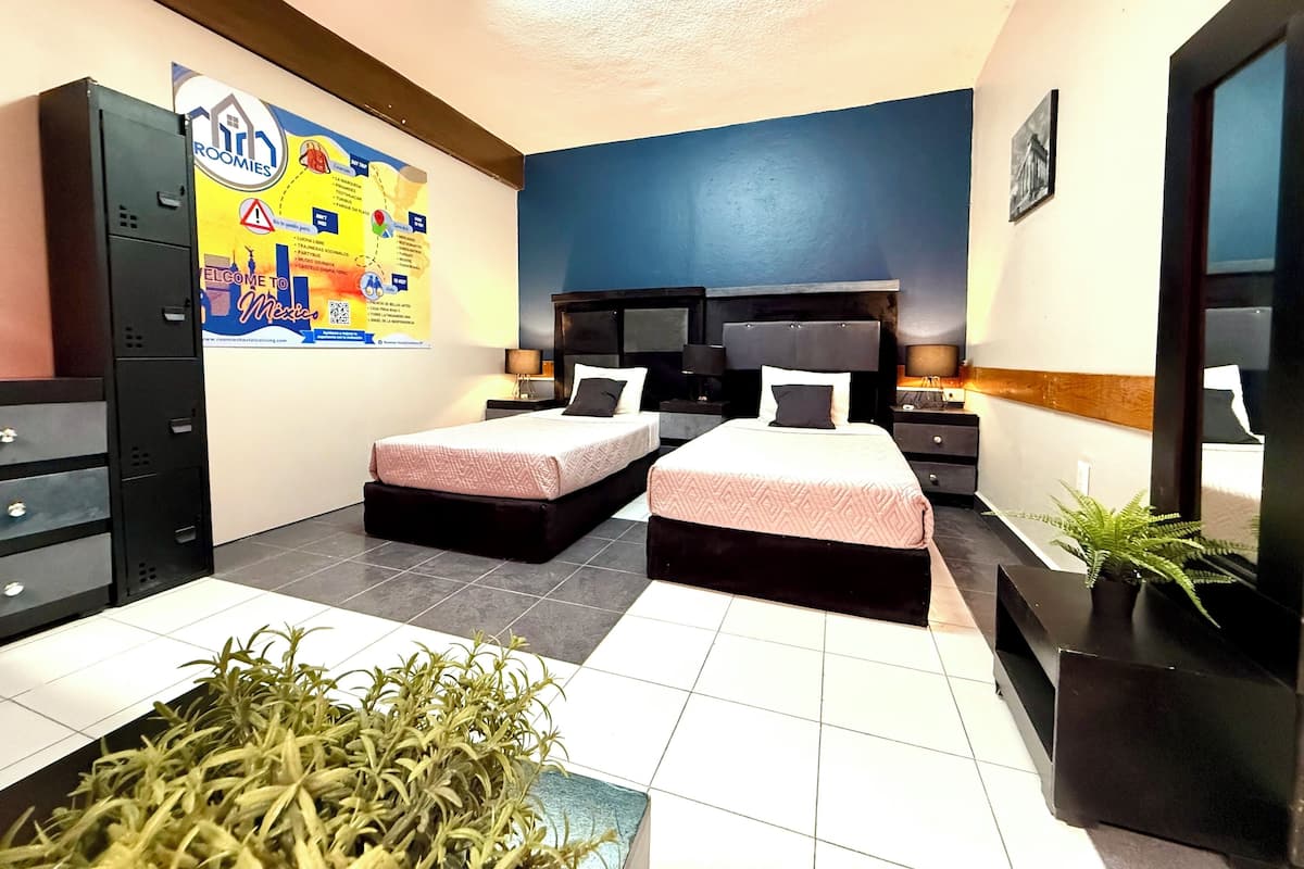 A hotel room with two beds, a desk, a chair, a plant, and a poster on the wall.