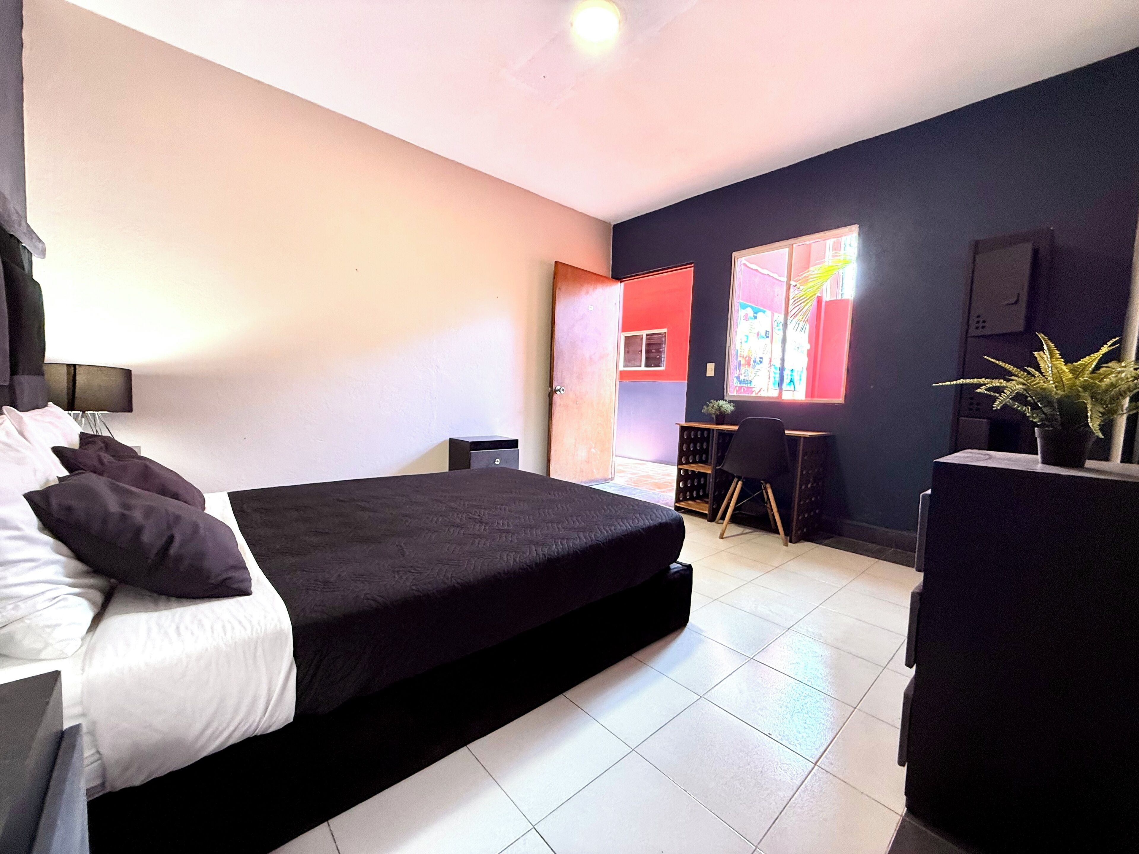 Camera moderna con un letto grande, una scrivania e una sedia. Si vede un tavolino con una pianta e una finestra con tende.